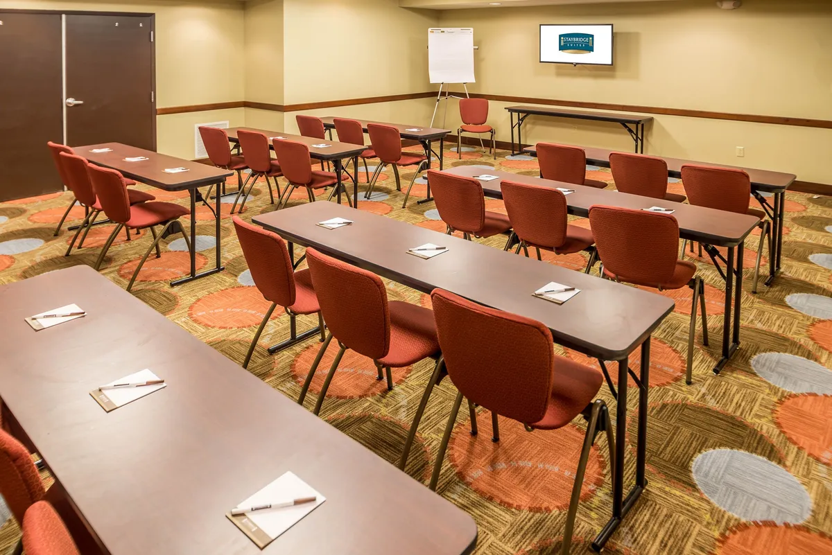 A spacious social/reception area arranged with rows of tables and vibrant red chairs, facing a presentation screen and whiteboard. The layout fosters collaboration and social interaction, perfect for evening guest receptions or informal gatherings.