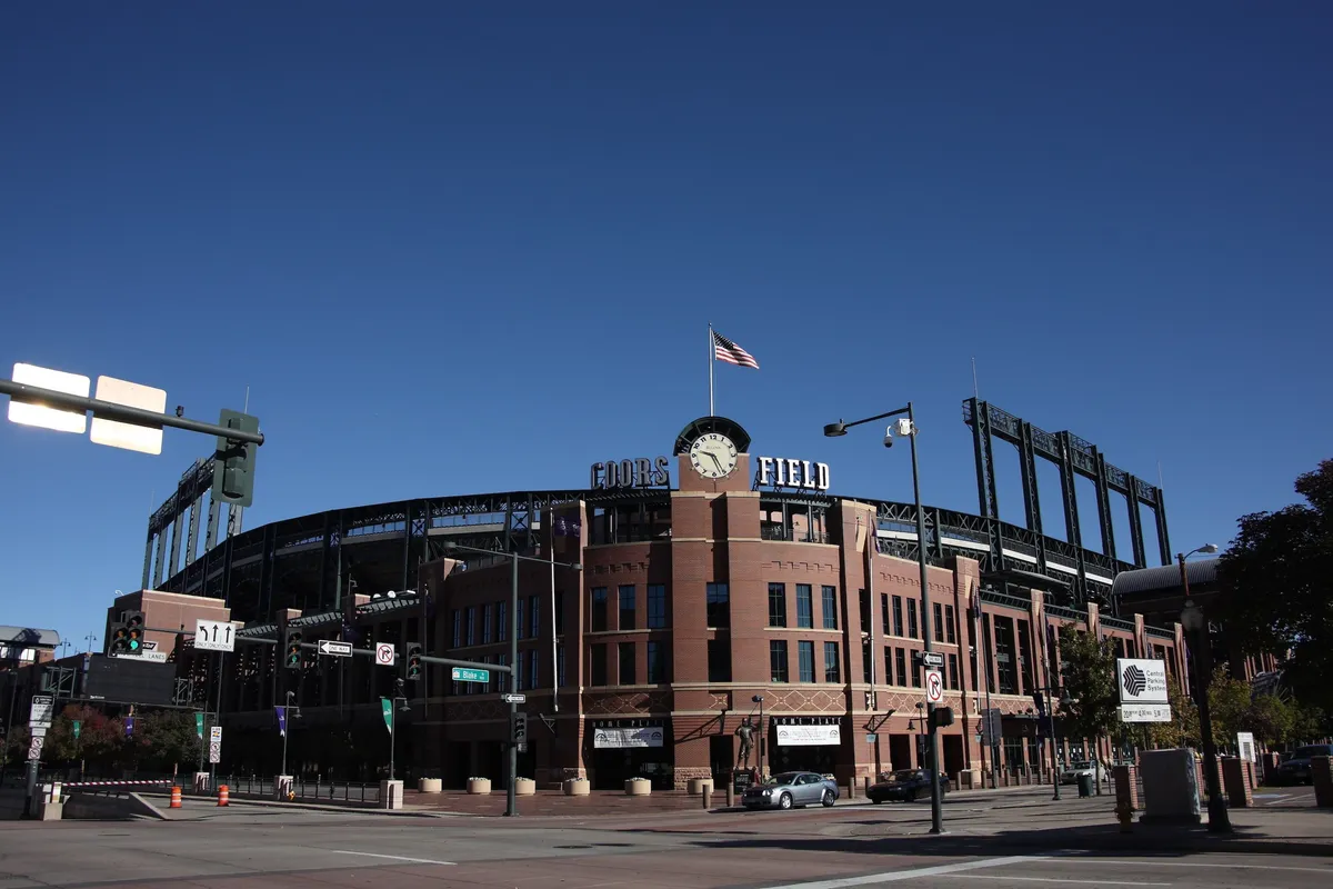 The image highlights the large brick facade of Coors Field, with a prominent clock and an American flag waving at the top. The atmosphere teems with anticipation, representing the thrill of live baseball games and community events.