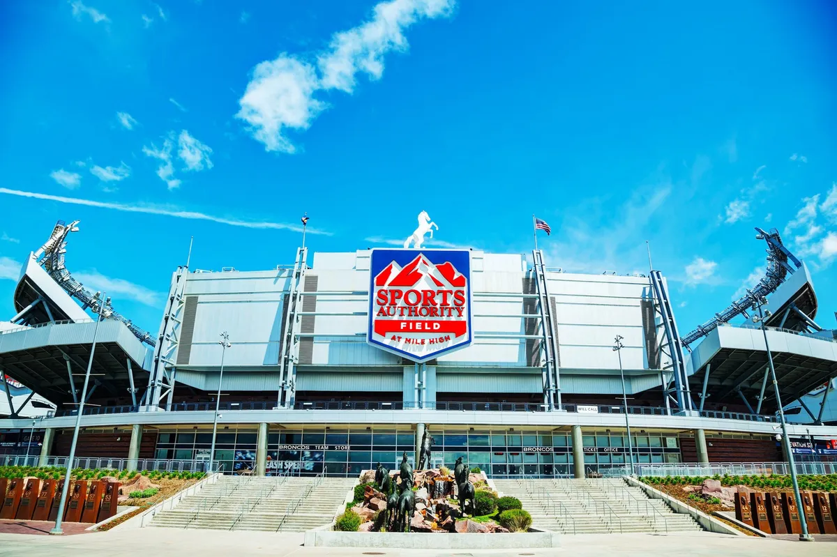 This exterior image features the impressive facade of a stadium, adorned with a large sign and surrounded by a lively landscape. The scene buzzes with the energy of fans gathering for a sports event or concert, capturing the excitement and vibrancy of Denver's entertainment offerings.