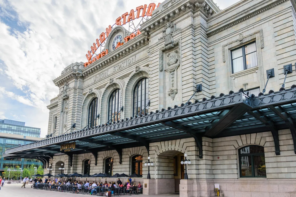 This image captures the grand facade of Union Station in Denver, with its iconic sign, inviting outdoor seating areas, and a lively mix of people. The scene radiates a sense of urban connectivity, vibrancy, and history, making it ideal for business travelers and networkers who value proximity to transport, dining, and city life.