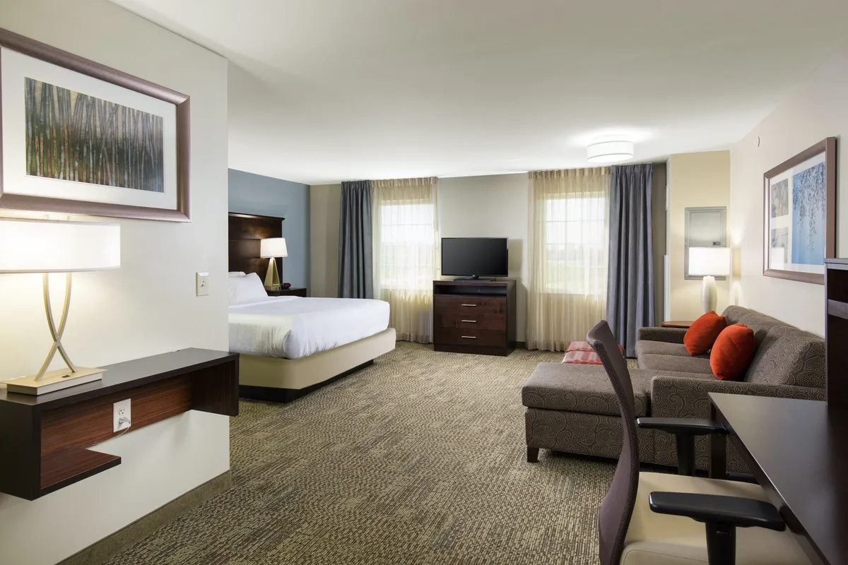 The image showcases a welcoming hotel suite interior featuring a plush queen-sized bed dressed in crisp linens, a cozy sofa adorned with accent pillows, a modern television perfect for movie nights, and tasteful framed artwork. A nightstand with a warm lamp adds to the inviting, home-away-from-home atmosphere, ideal for relaxing, reading, or catching up on work.