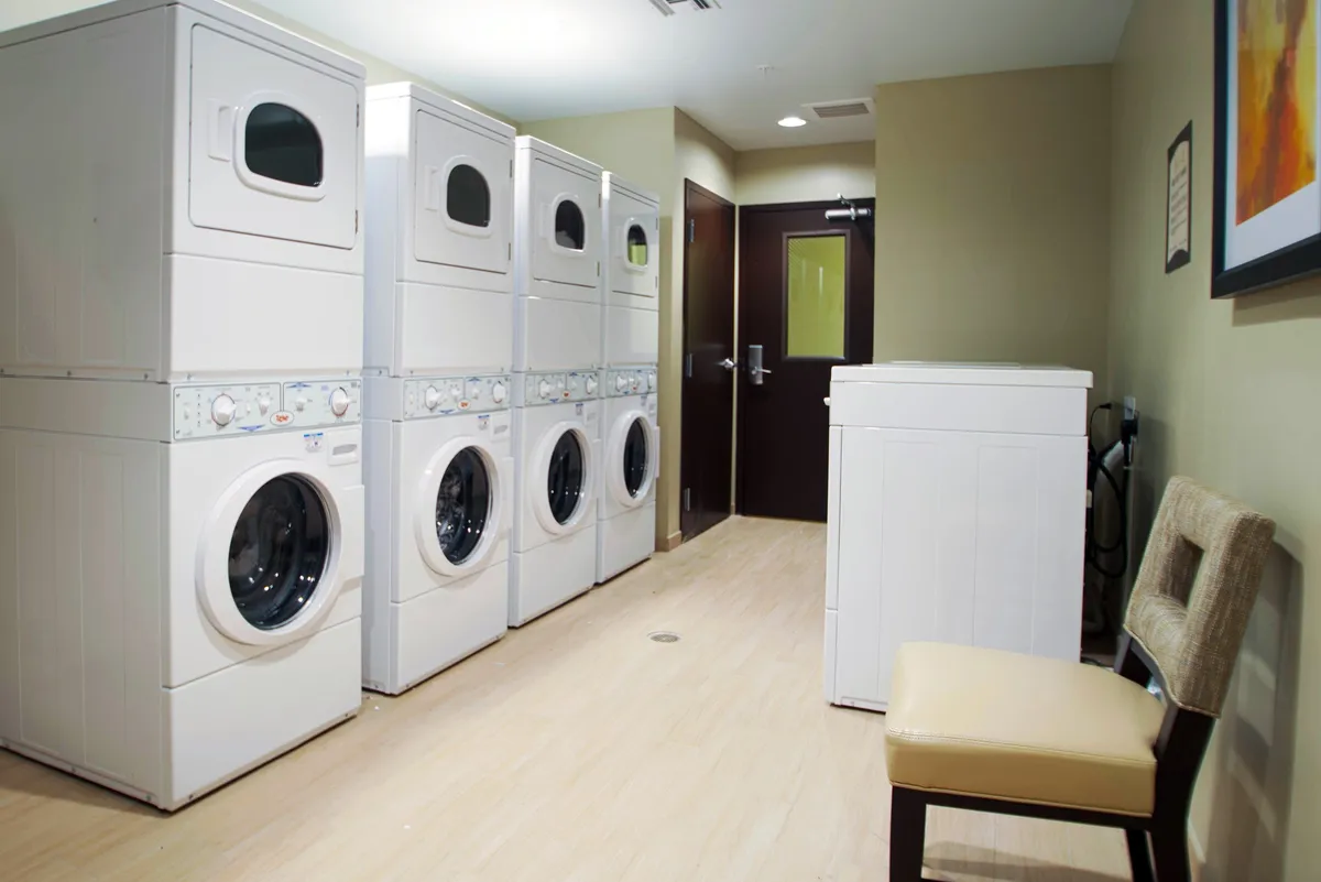 A clean, practical laundry facility featuring stacked washer and dryer units, a single washing machine, and a comfortable chair for guests waiting for their laundry. The setting is bright and organized, emphasizing ease of use and convenience.