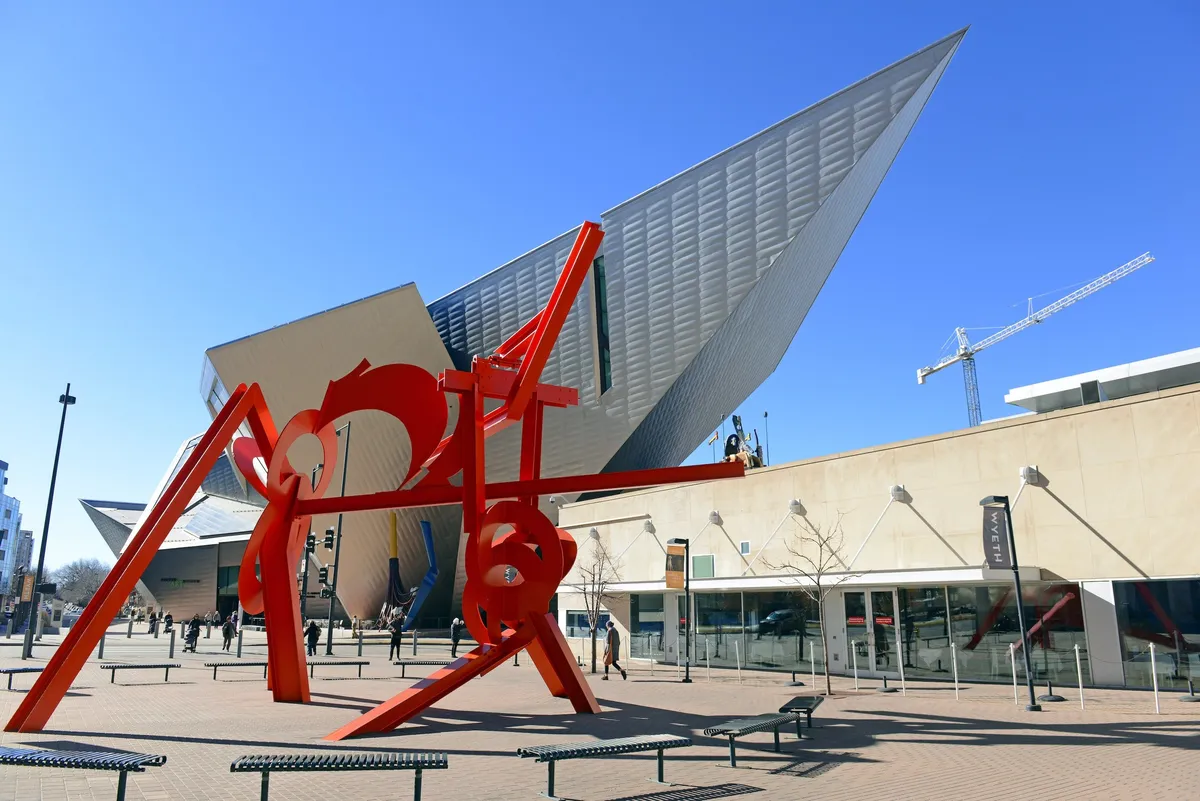 A large, striking red sculpture sits in front of a modern architectural building, with benches and open gathering spaces in the foreground. The dynamic setting suggests opportunities for public gatherings, art appreciation, and relaxed networking in an urban context.